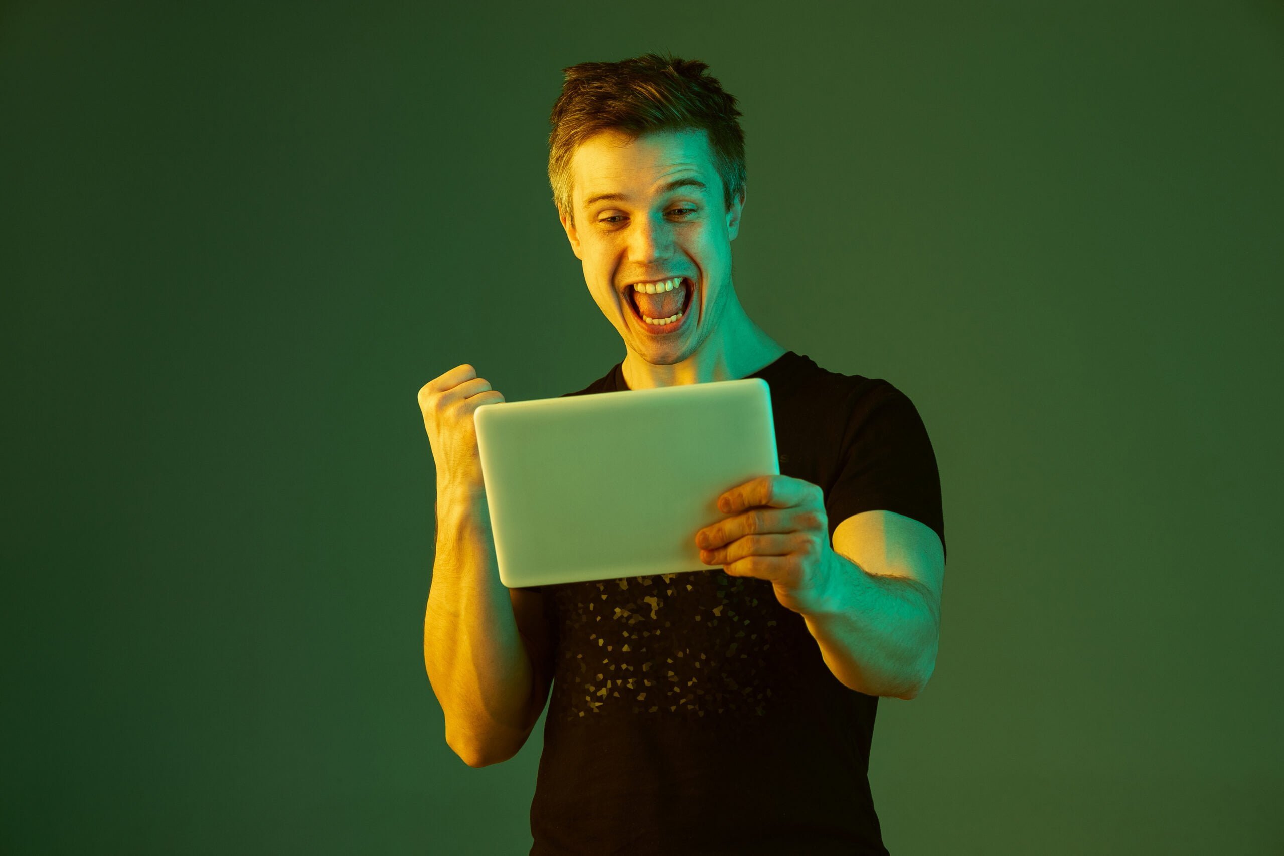 holding tablet celebrating win bet game caucasian man s portrait green studio background neon light beautiful male model concept human emotions facial expression sales ad scaled 1